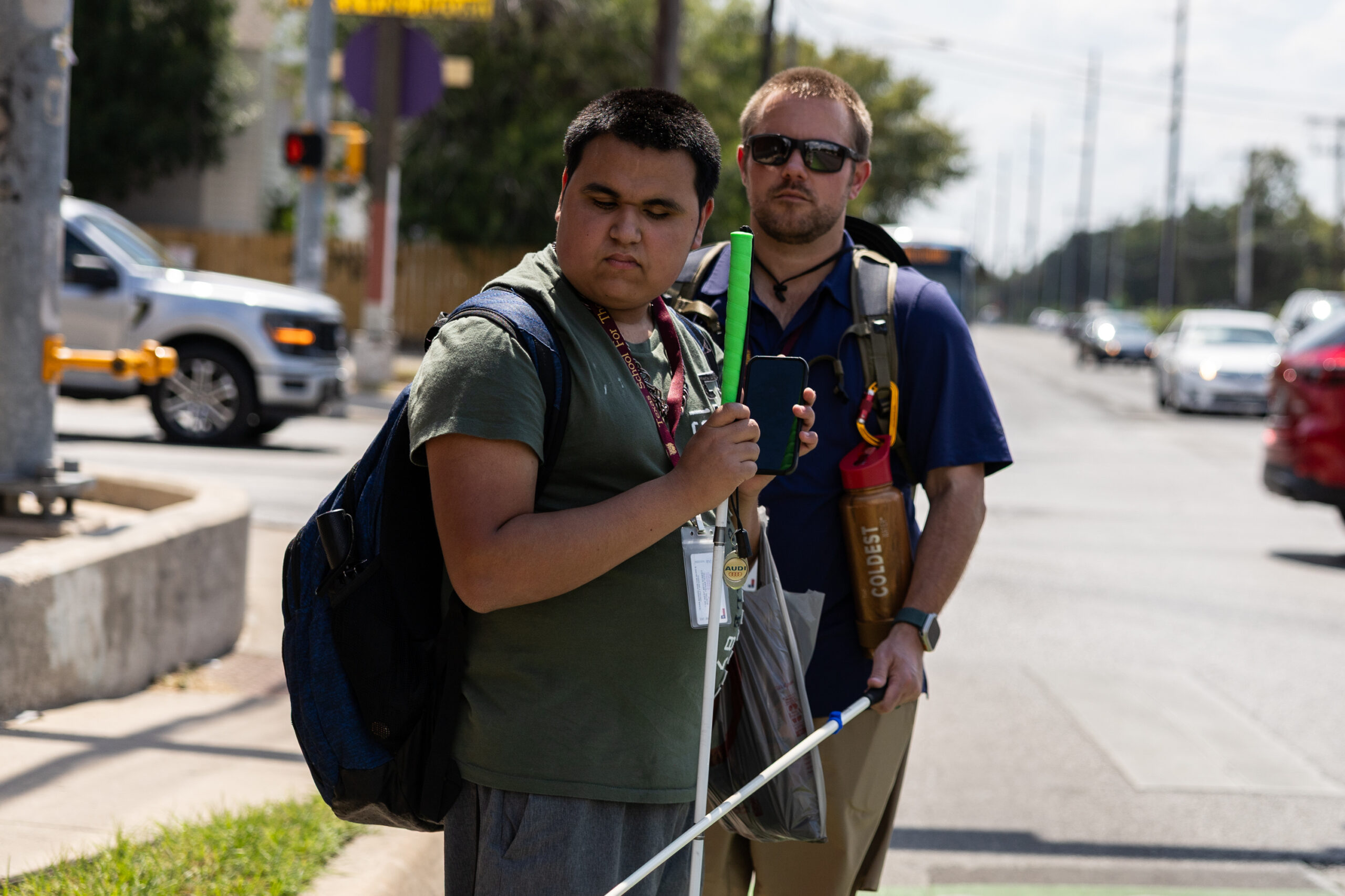 Two men wait at a crosswalk holding white canes. This is Drew Bernet and Oscar Garcia.