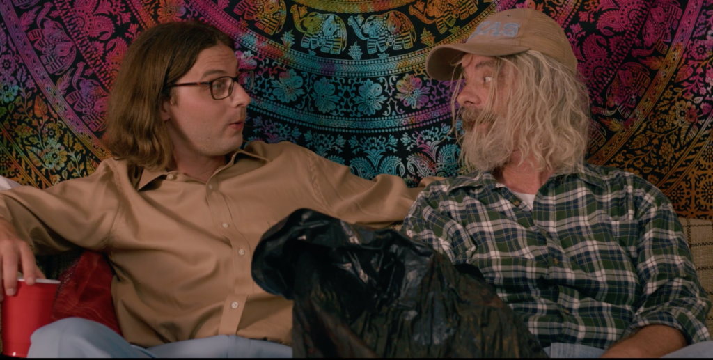 A close-up photo of a young man with glasses and an older man with unwashed hair sitting on a couch.