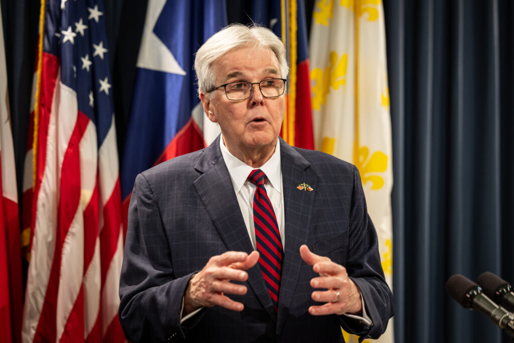 A man in a dark suit and red-and-navy striped tie gestures while speaking into microphones.