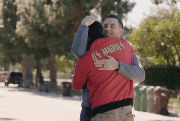 Two people hug tightly on a tree-lined street. One person is wearing a red jacked with the words "U.S. Marines" on the back.
