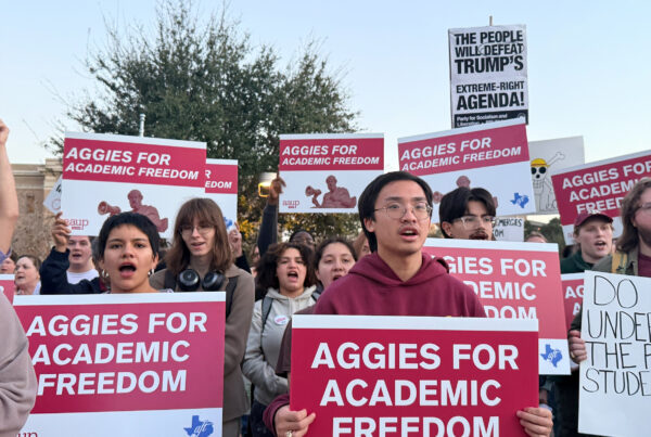Students in a group at an outdoor rally hold maroon and white signs reading "Aggies for Academic Freedom."