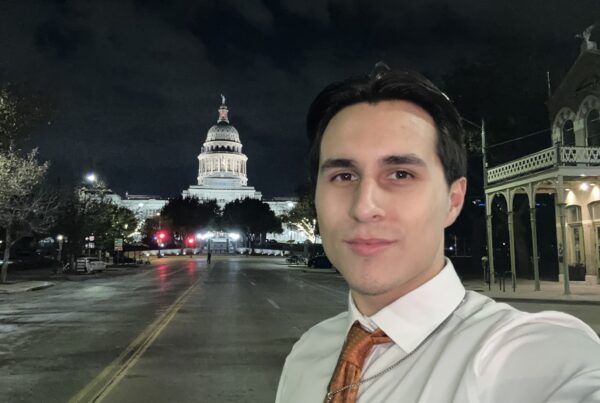 A person wearing a suit shirt and tie takes a selfie at night in front of the Texas Capitol building.