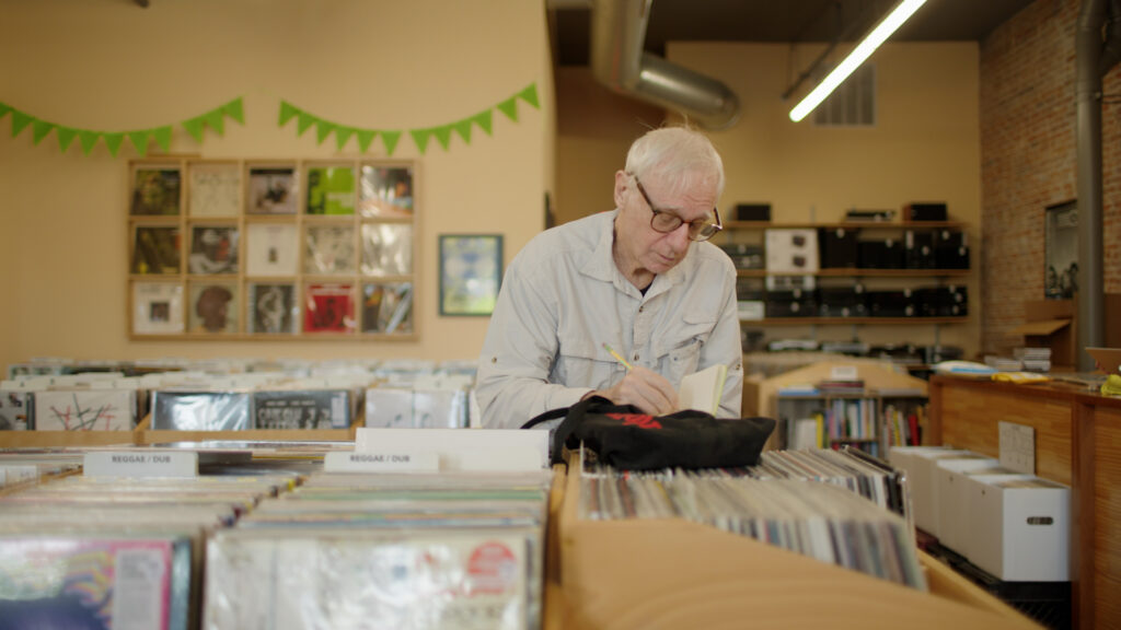 A still image from the film "The Last Critic" shows a man taking notes in a record store.
