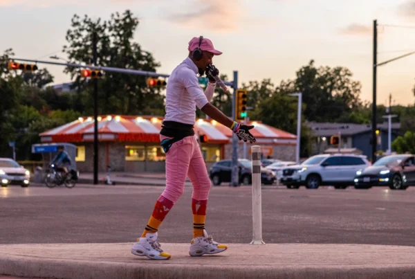 A man wearing bright athletic clothes, wearing headphones and holding a microphone is at a street corner as the sun sets.