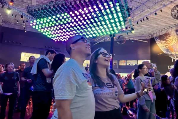 Two people wearing NASA shirts are front and center in a crowd watching a screen in awe. The room is lit with colorful lights.