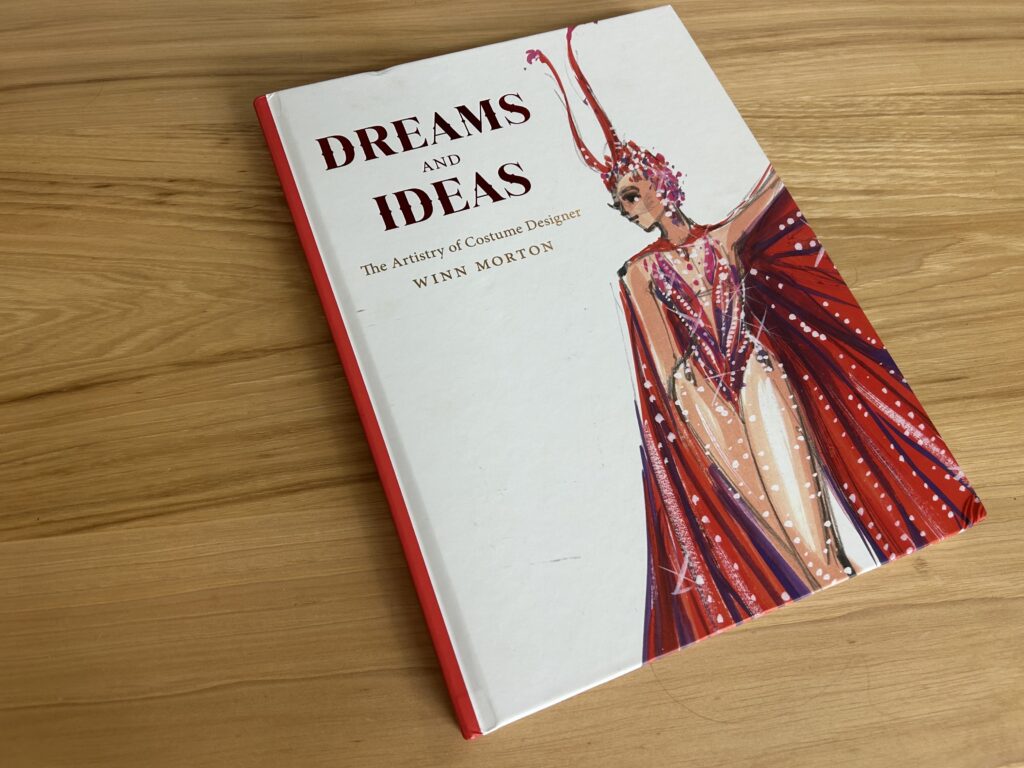 The book "Dreams and Ideas" is shown sitting on a wooden table. The cover shows a sketch of a show girl in a red leotard and cape.