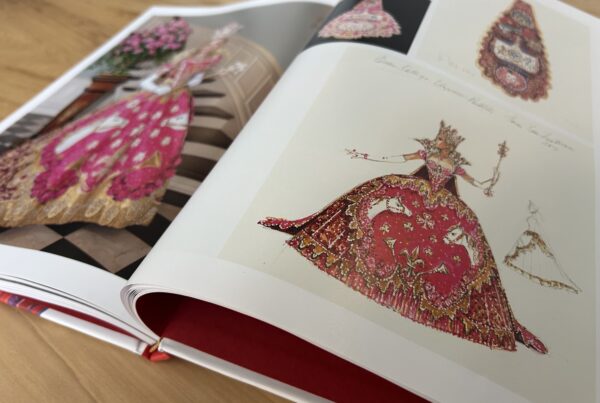 A close-up photo of an open book shows the sketch of an elaborate pink gown on one page and a photo of a woman in the same gown on the other page.
