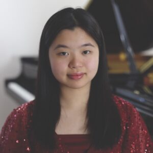 A headshot of a young woman with long hair wearing a red dress and sitting in front of a piano.