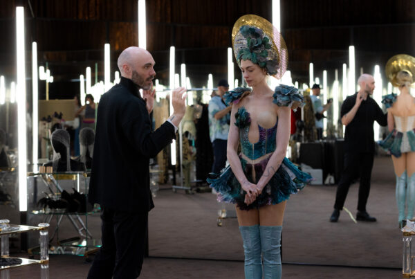 A behind-the-scenes photo during the making of "Mother Mary." Shows Lowery, dressed all in black, speaking to Hathaway, dressed in a short showgirl's costume, while on a set with lots of vertical lighting.