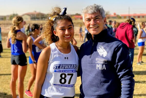 Two people smile for the camera. They are outdoors on a sunny day. Mia is wearing a racing bib and uniform and other runners are seen behind the pair.