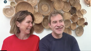 The couple smile at one another as they sit in front of a wall covered with baskets of various sizes.