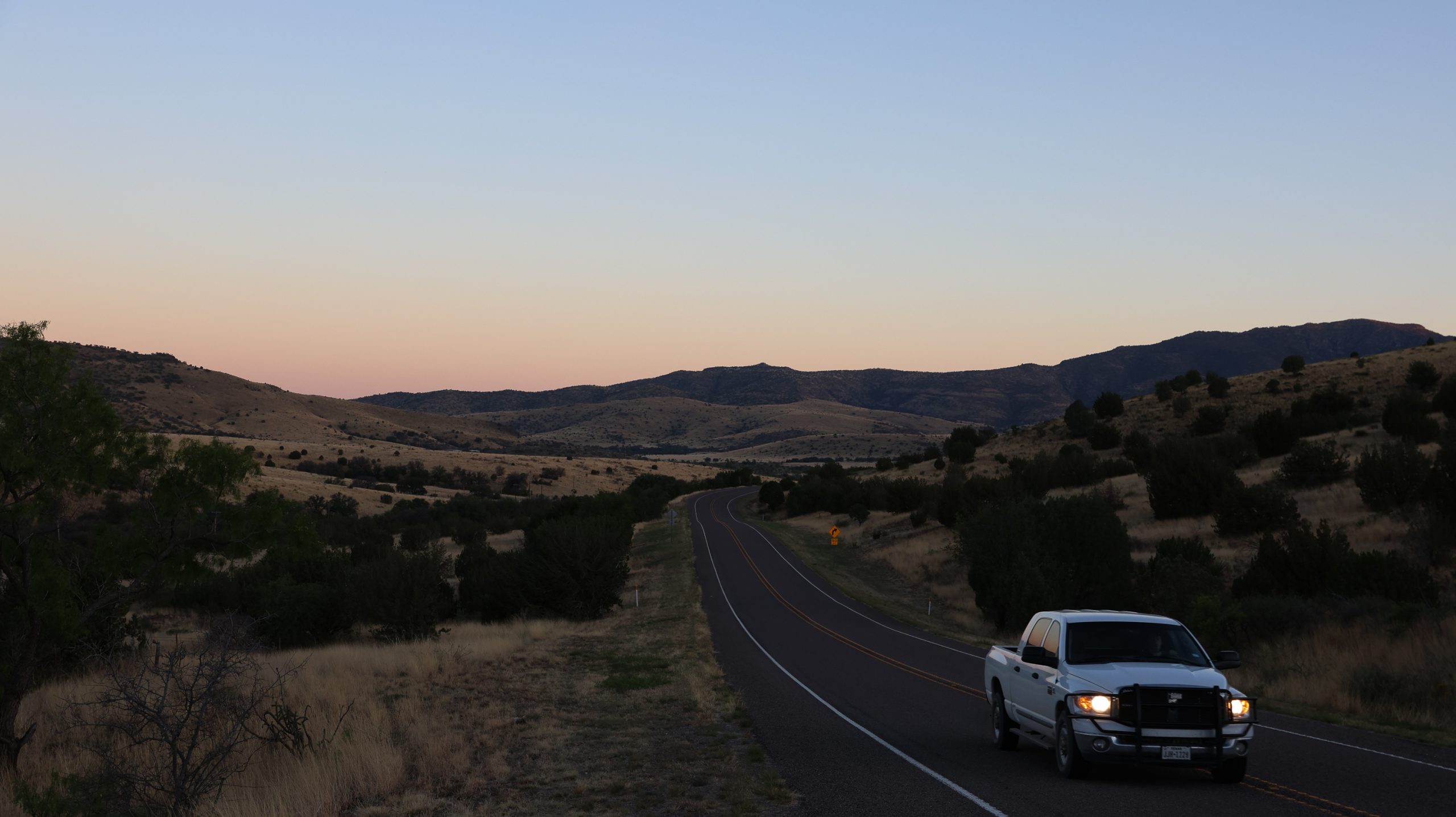 A truck is seen driving in the foreground on a road with a range of mountains seen in the distance as the sun sets.