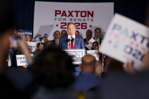 Ken Paxton stands at a podium in front of a crowd of supporters at a rally during his Senate campaign. He is wearing a blue suit and a red tie and addressing the crowd. Behind him, a white and red Paxton for Senate 2026 banner takes up much of the stage
