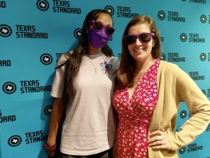 Two people pose for a photo in front of a blue background with the Texas Standard logo on it.