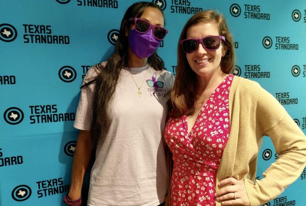 Two people pose for a photo in front of a blue background with the Texas Standard logo on it.