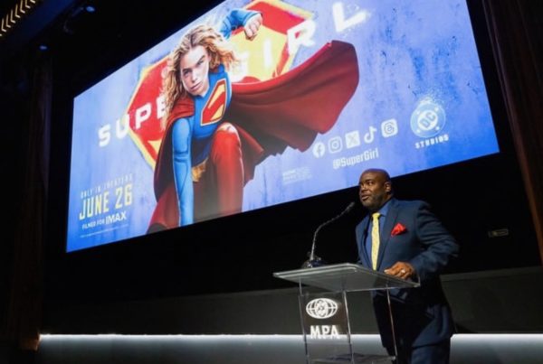 A wide shot of a man in a suit behind a clear podium with the MPA logo on it. Behind him is a wide screen with the "Supergirl" poster on it.