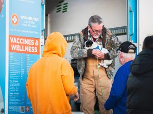 A man carries a small dog outside the back of a bus. Next to him is a sign reading "vaccines & wellness."