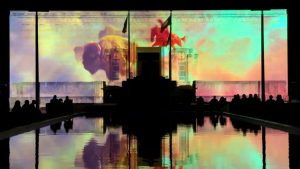 A projection of a buffalo and a person riding a horse is colorful on the side of a rectangular building. People watch and the image is reflected in a pool.