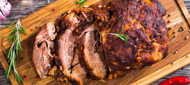 Sticky-Sweet Grilled Pork Shoulder – Memorial Day Backyard BBQ Series Oven-Barbecued Pork shoulder, cut on slices on chopping board with rosemary, peppercorn and bay leaf on old dark planks with garlic and chili on background, view from above, close-up