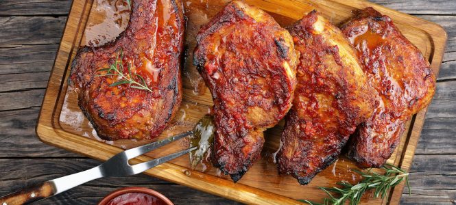 Orange-Ginger Pork Chops – Harvest Grilling Series Roasted tender, juicy Pork Chops with bay leaves, rosemary, spices on a wooden cutting board on a rustic table with barbecue sauce and fork, easy recipe for Christmas dinner, vertical view from above