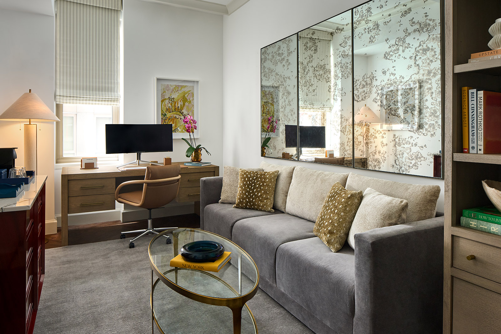 A wide view of a hotel suite with a gray sofa, wooden desk, bookshelf, and large mirror.