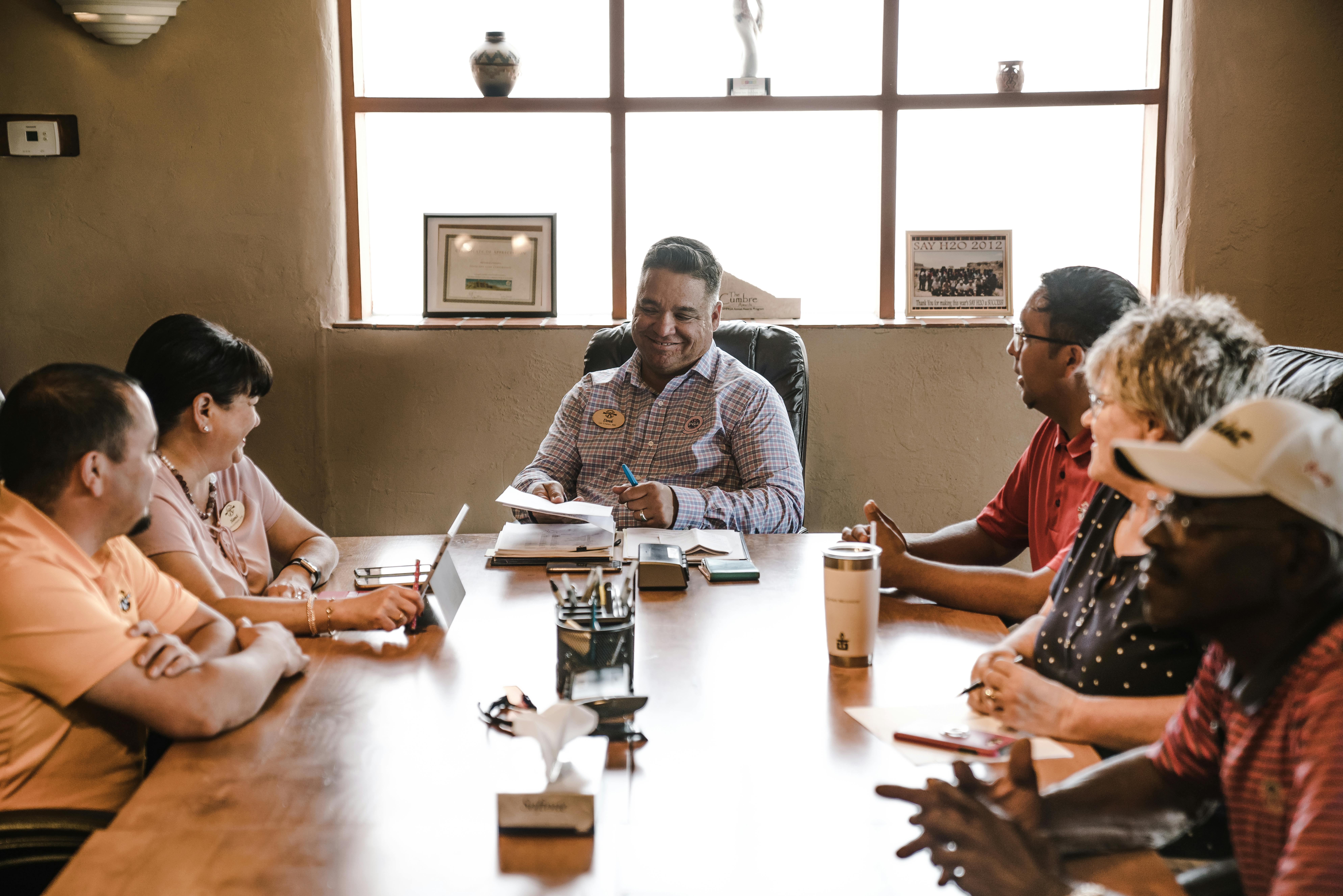 A diverse group of people sit around a conference table engaged in a friendly and productive meeting, with one person leading the discussion and smiling.