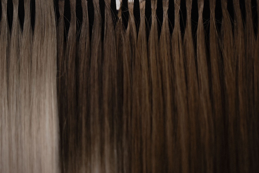A lineup of different colored hand-tied hair extension wefts hanging against a black background.