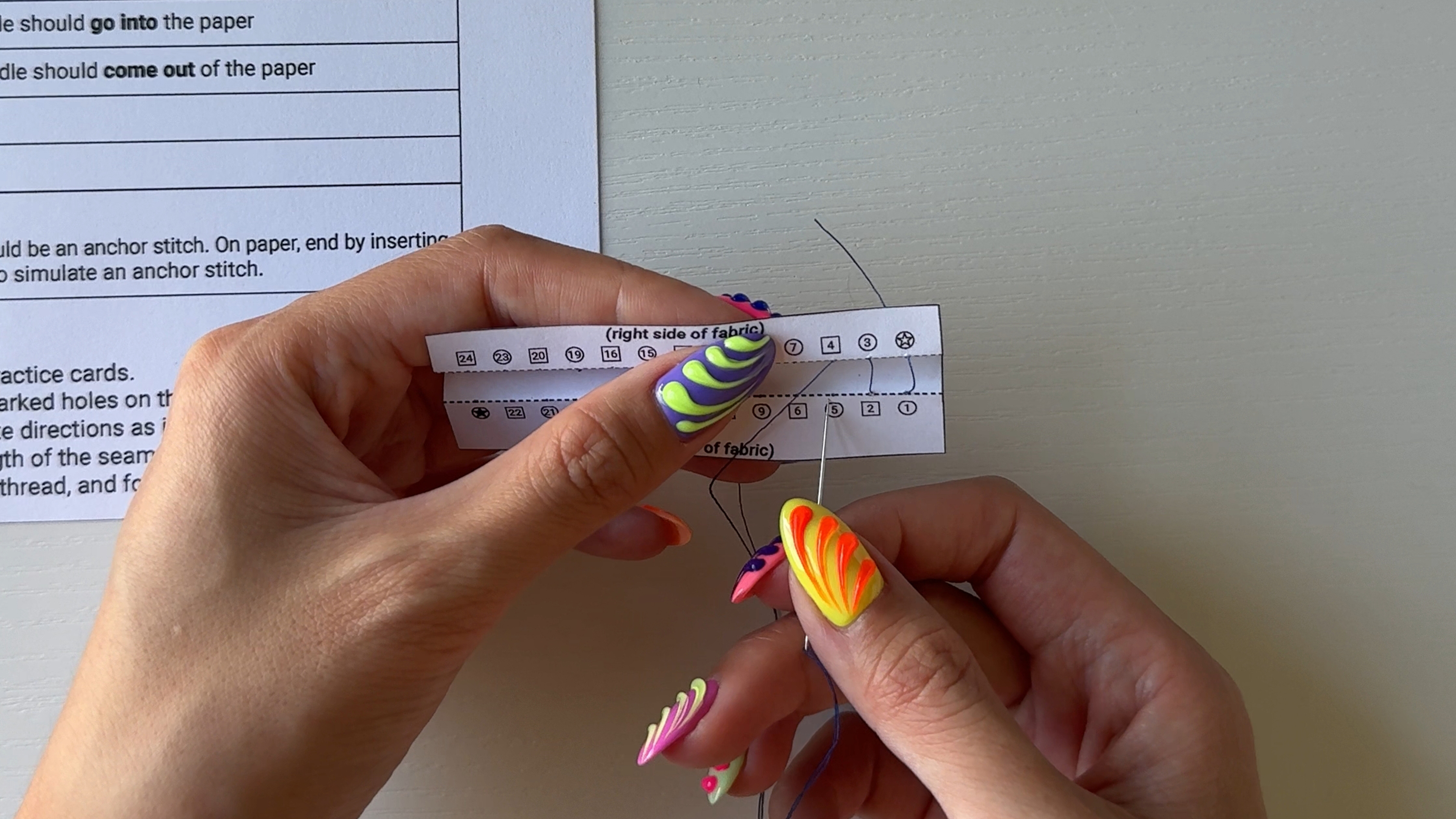 Up-close of two hands hand sewing a paper printout included in the online course.