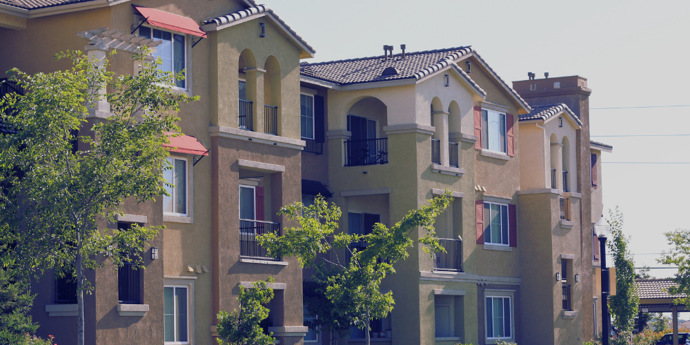Three story apartment building. Two-toned tans stucco facade and terra cotta tile roof. Pretty trees.