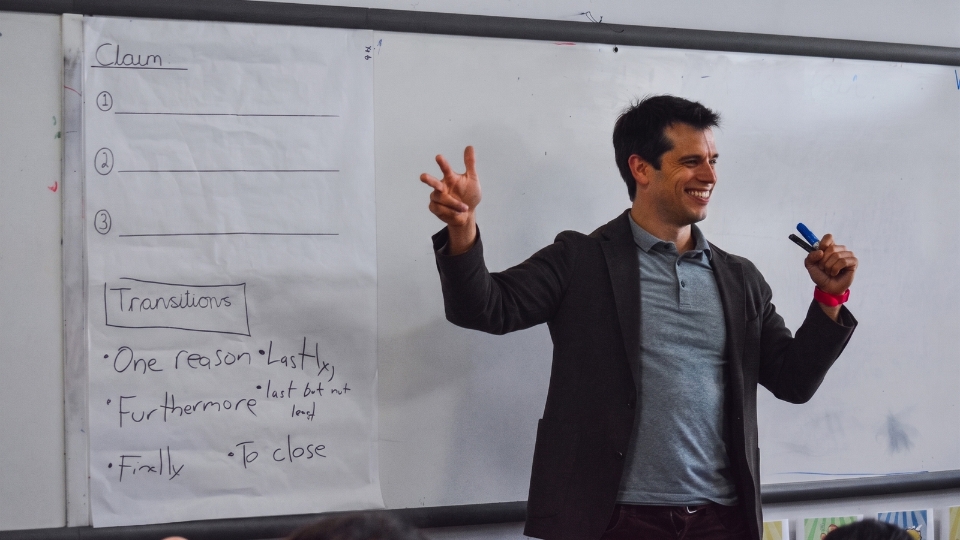 Gerrit teaches at the front of a classroom, smiling and gesturing beside an anchor chart of claims and transition words.
