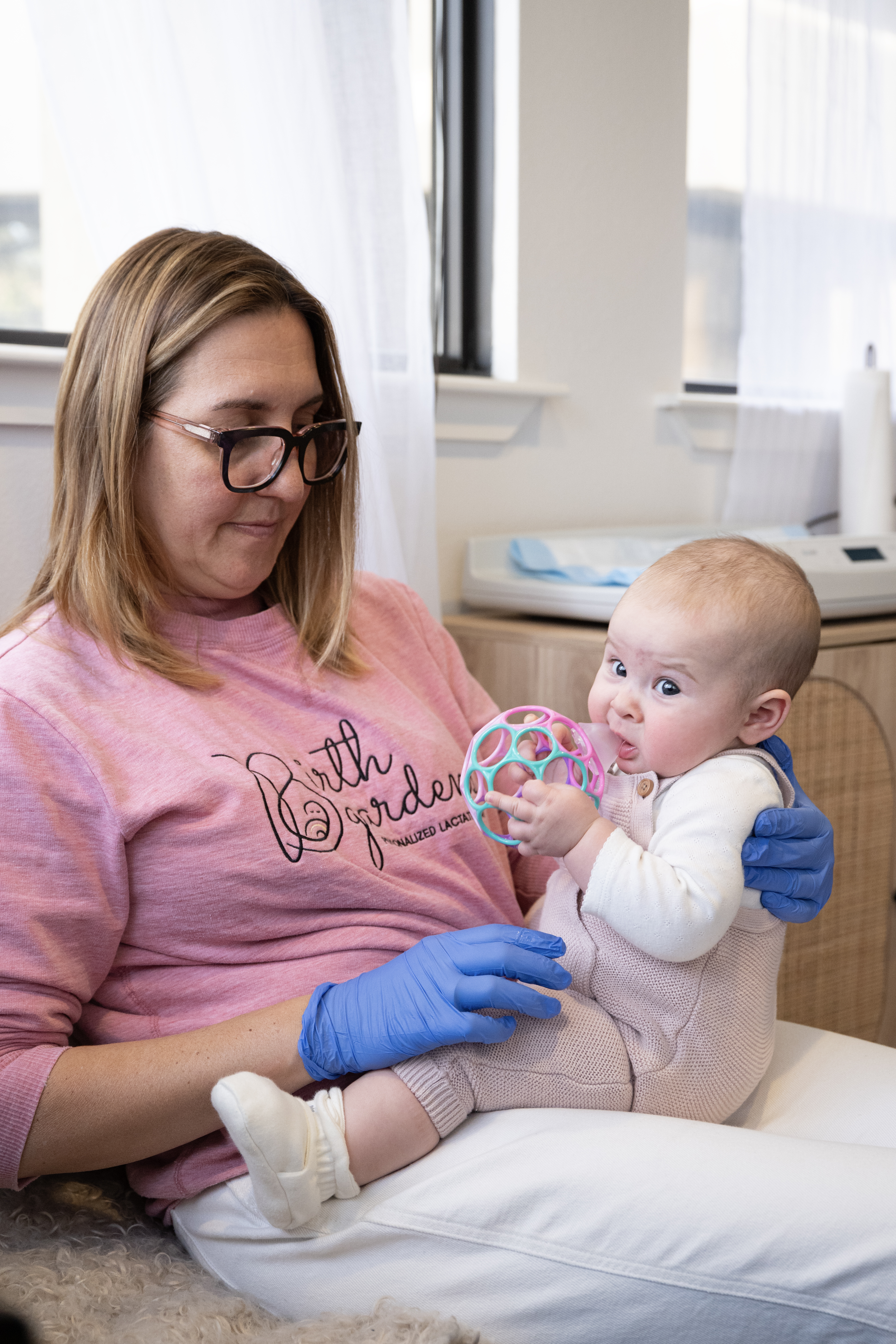 Lactation consultant wearing a Birth Garden LACTATION sweatshirt and medical gloves supporting an infant who is sitting on her lap and holding a colorful toy during a feeding or postpartum care session.