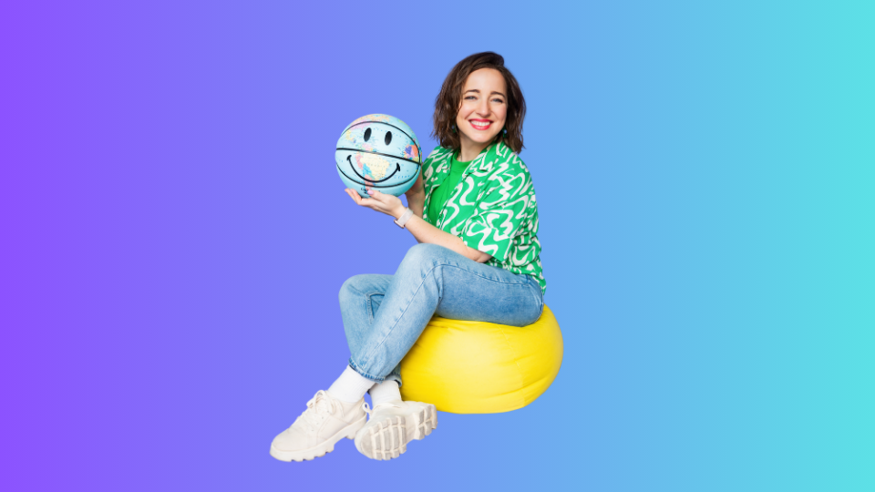 Image shows the course creator Agnese Spona sitting on the cushion and holding a basketball painted like a globe and with smiley face on top of it. 