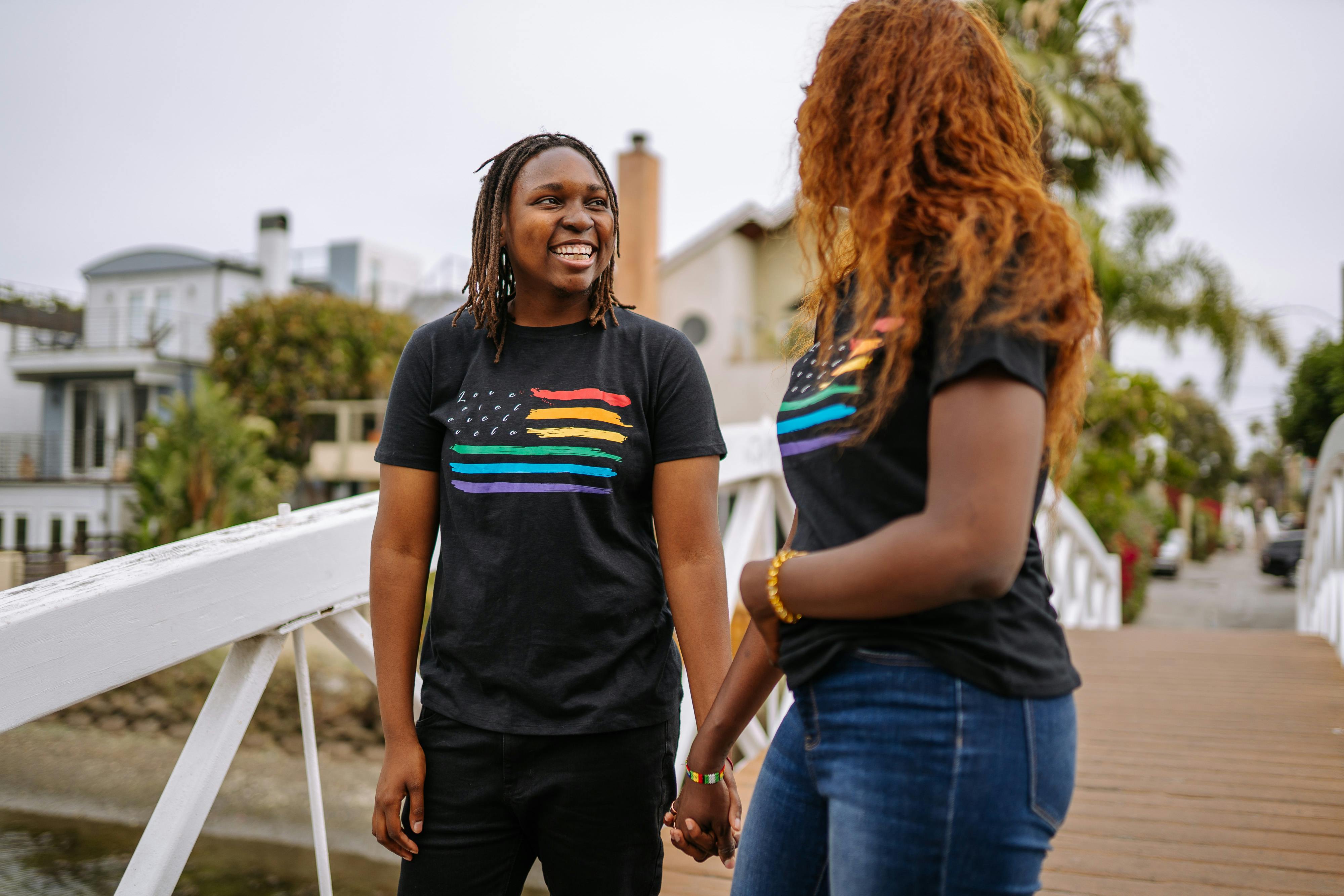 Two dark skinned people are seen engaging in a joyful conversation. Both are wearing black t-shirts with an american pride flag on them
