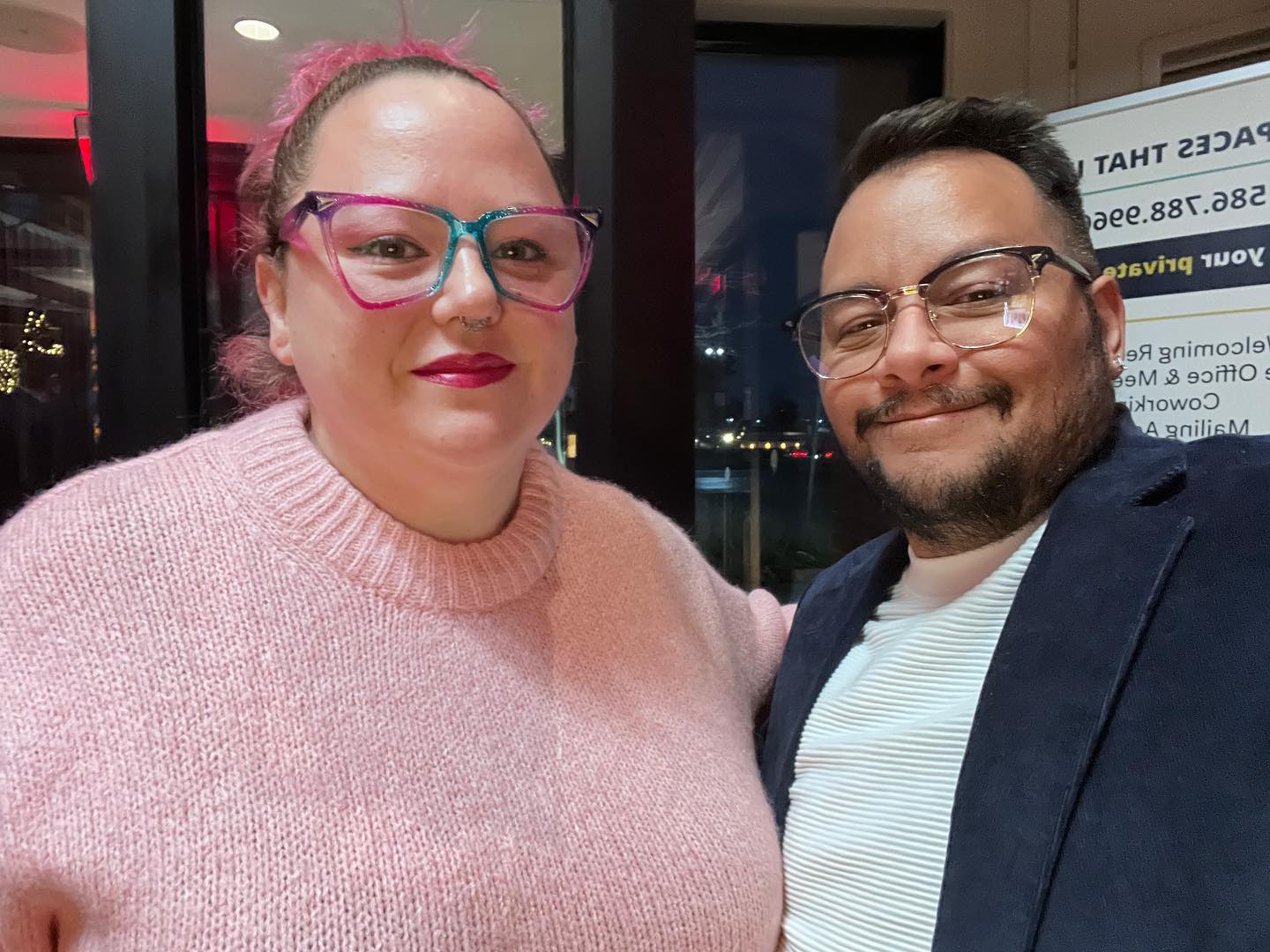 Heather and Jack Knoxville are seen smiling at the camera. Heather is a white woman wearing a pink sweater with multicolored glasses. Jack is a melanated trans man wearing a navy blue jacked over a white long sleeve shirt also wearing glasses.
