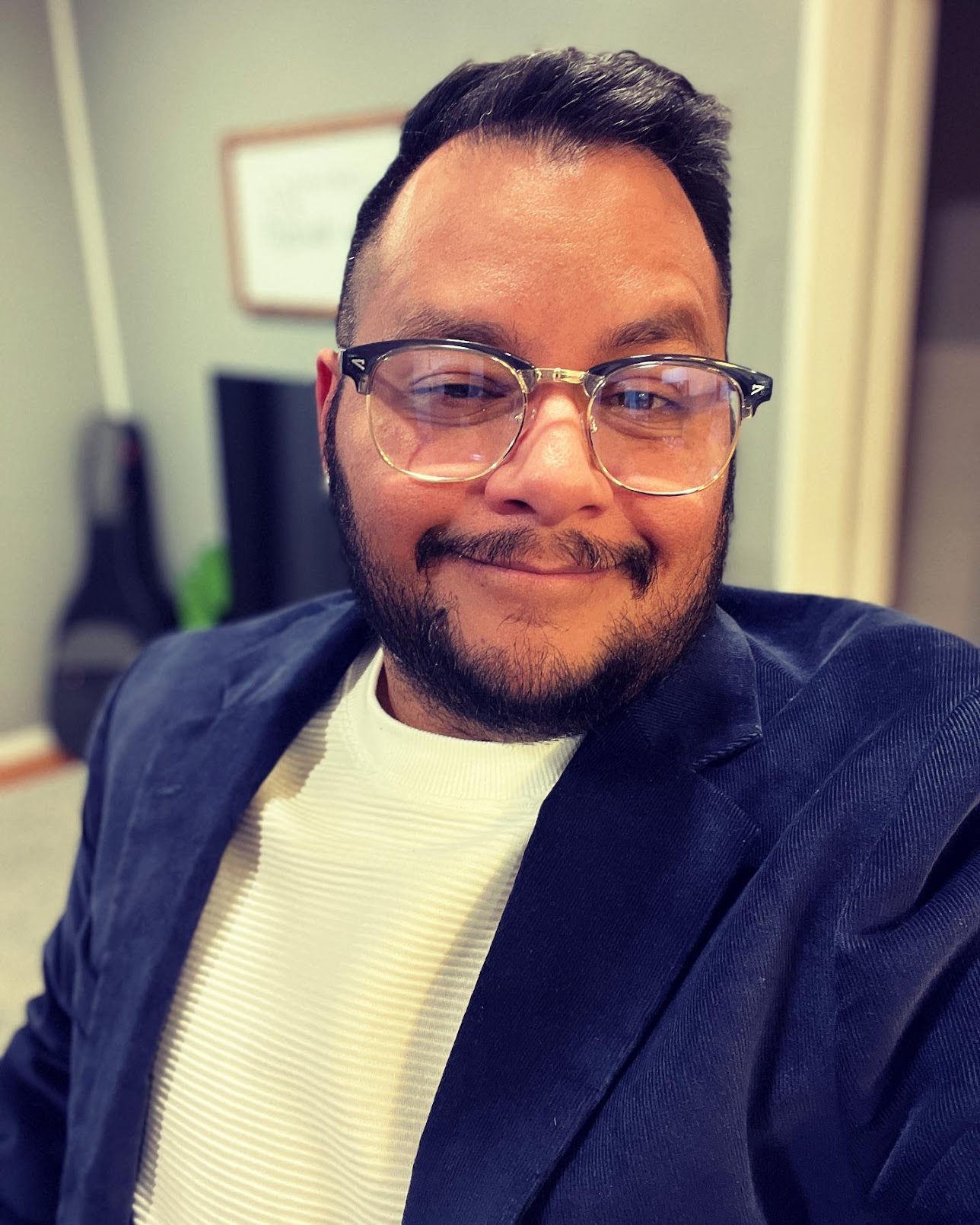 Jack is a melenated afro-latino trans man seen wearing a white shirt and blue blazer. He has short dark brown hair and has a neatly trimmed mustache and beard