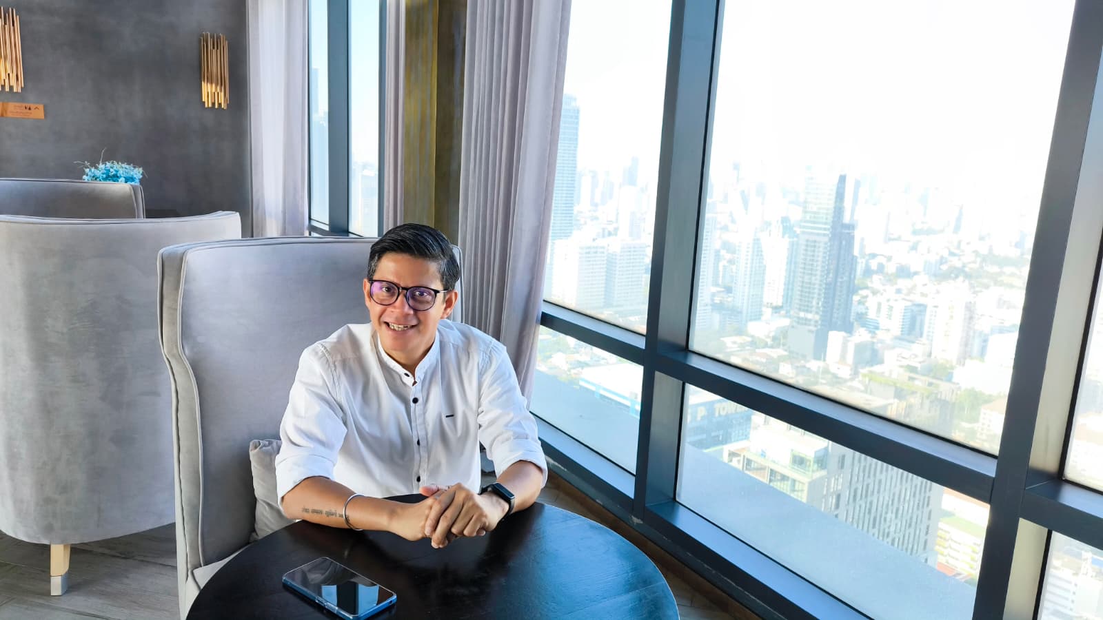 men sitting in an armchair resting his hands at the table, in the background is window with the view overlooking a big city 