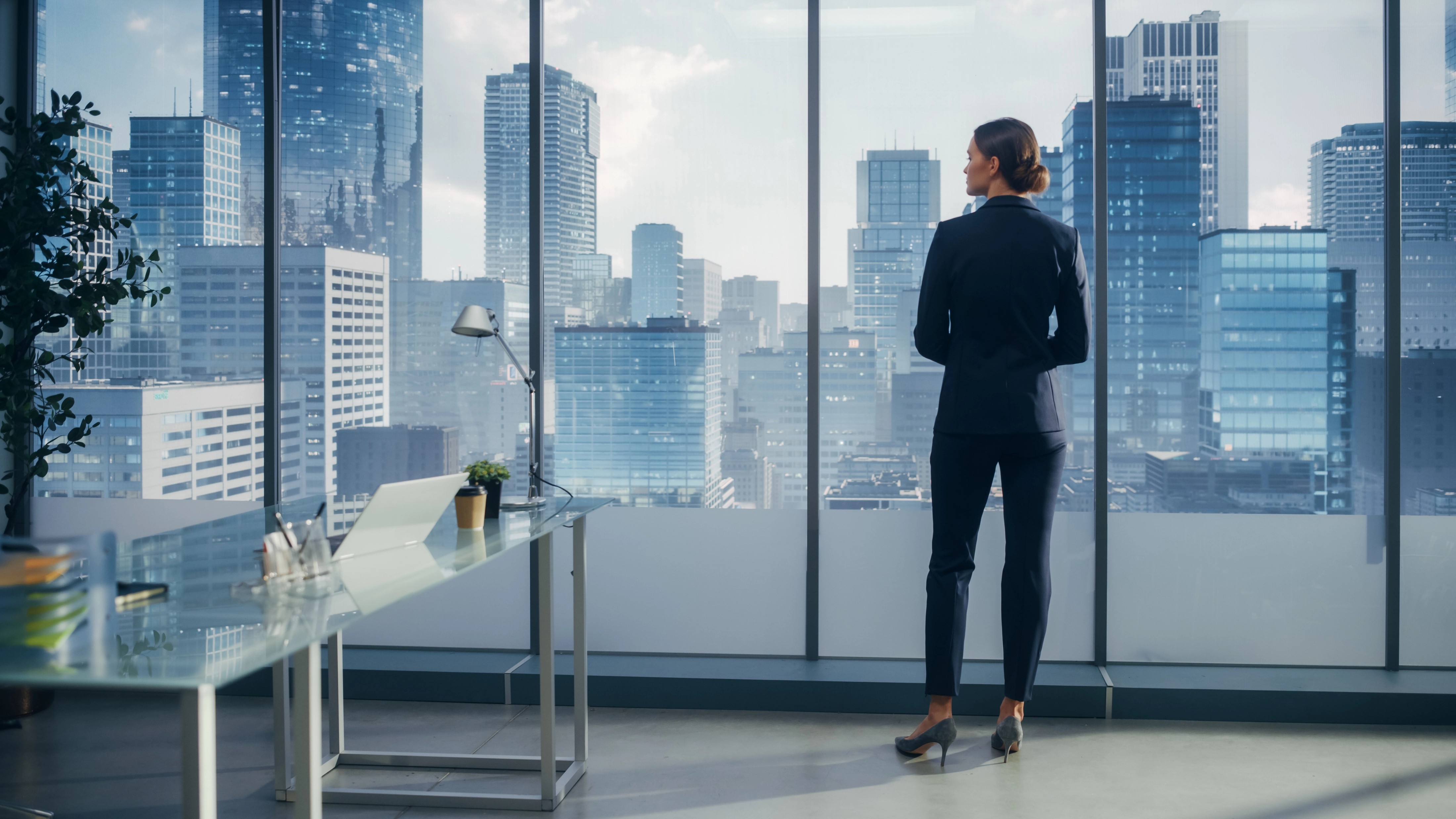 A woman in a high-rise office looking out the window onto other tall buildings in a city.