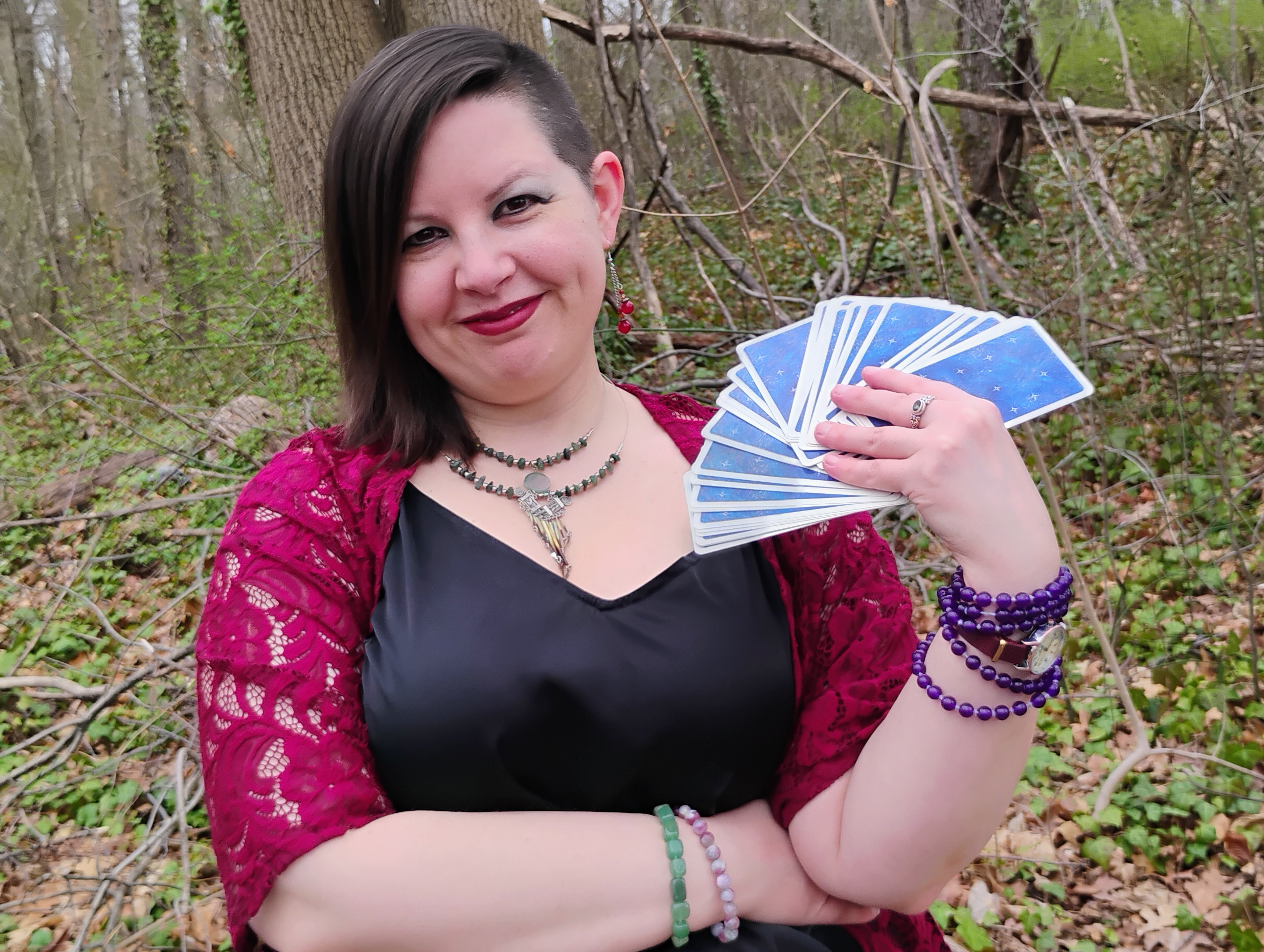 Photograph of Evylyn Rose, Empowerment Guide and Tarot Reader; she is outside, in a wooded area, holding a fanned deck of Tarot cards with a smile on her face.