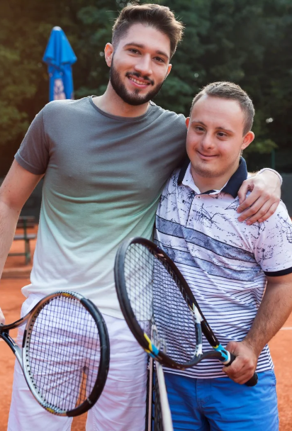 Two men, one with down syndrome holding tennis rackets. Man without down syndrome has arm over other mans shoulder.