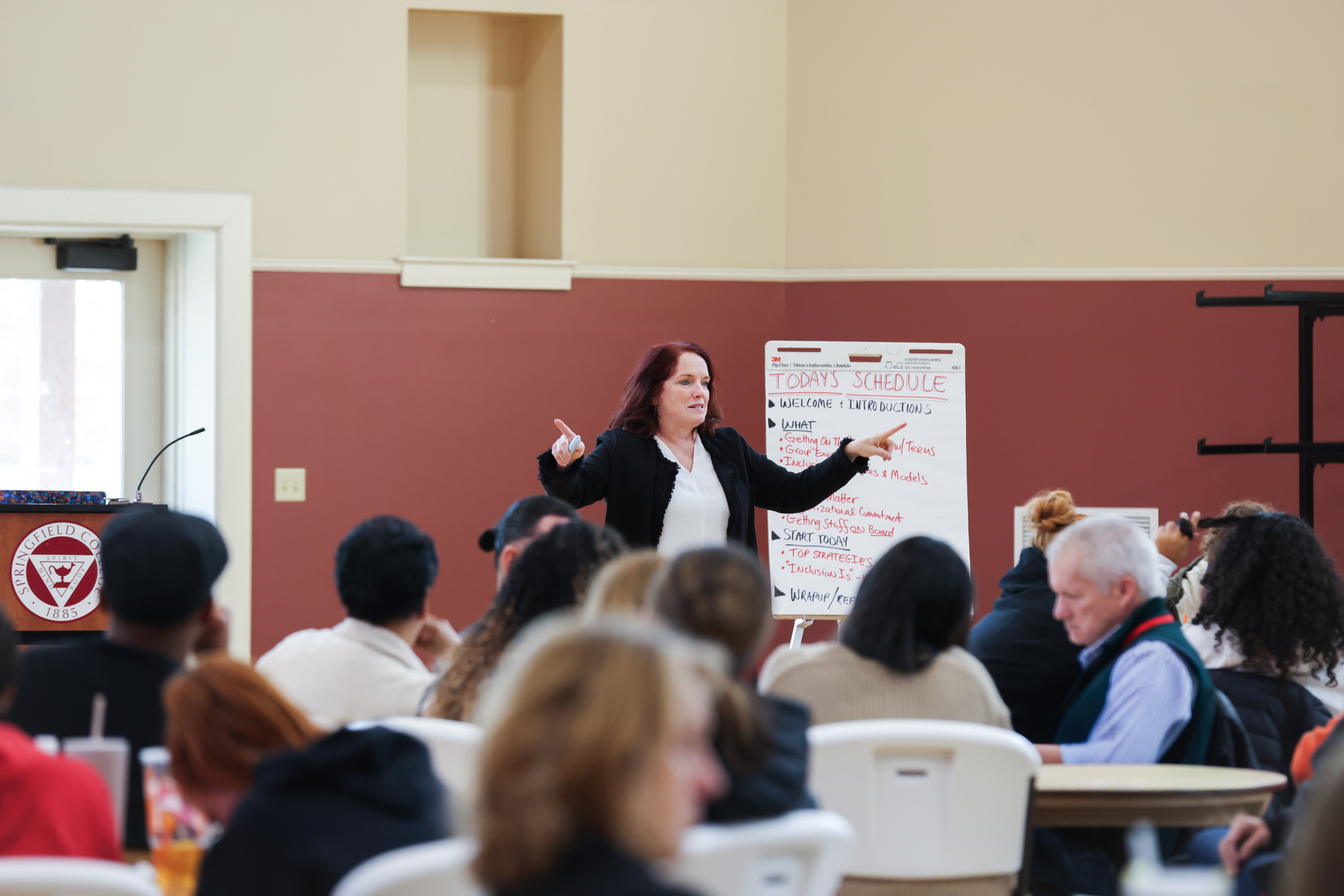 Woman presenting in front of crowd, back of crowd heads face camera
