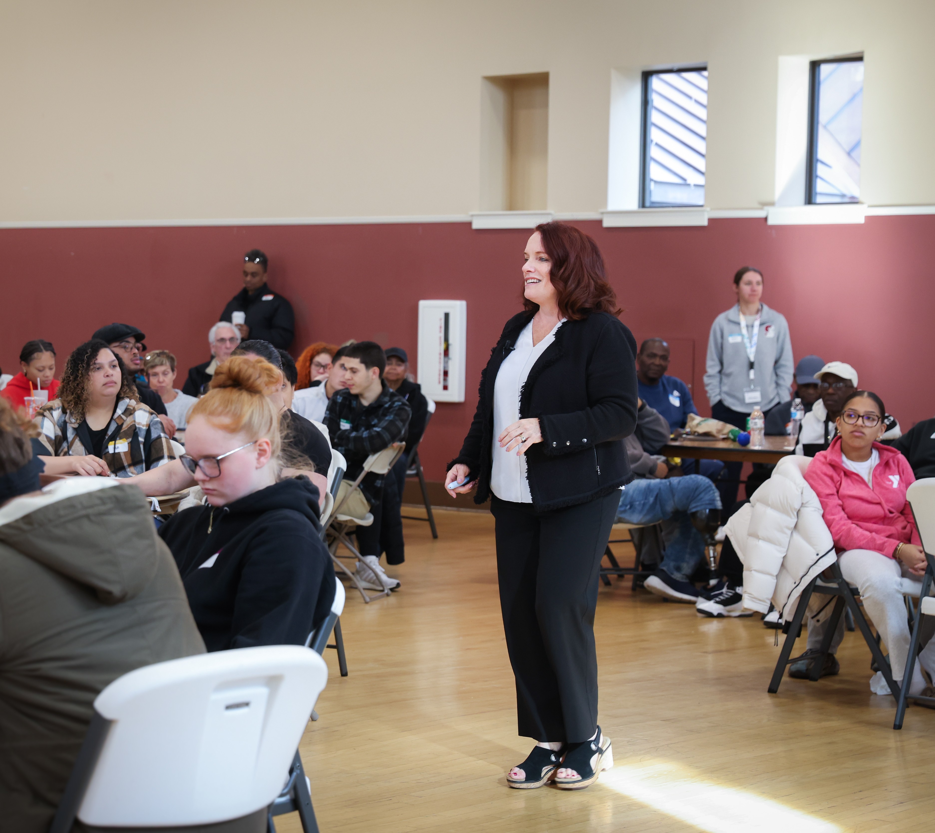 woman in black clothing smiling and walking around a room filled with people sitting at circular tables