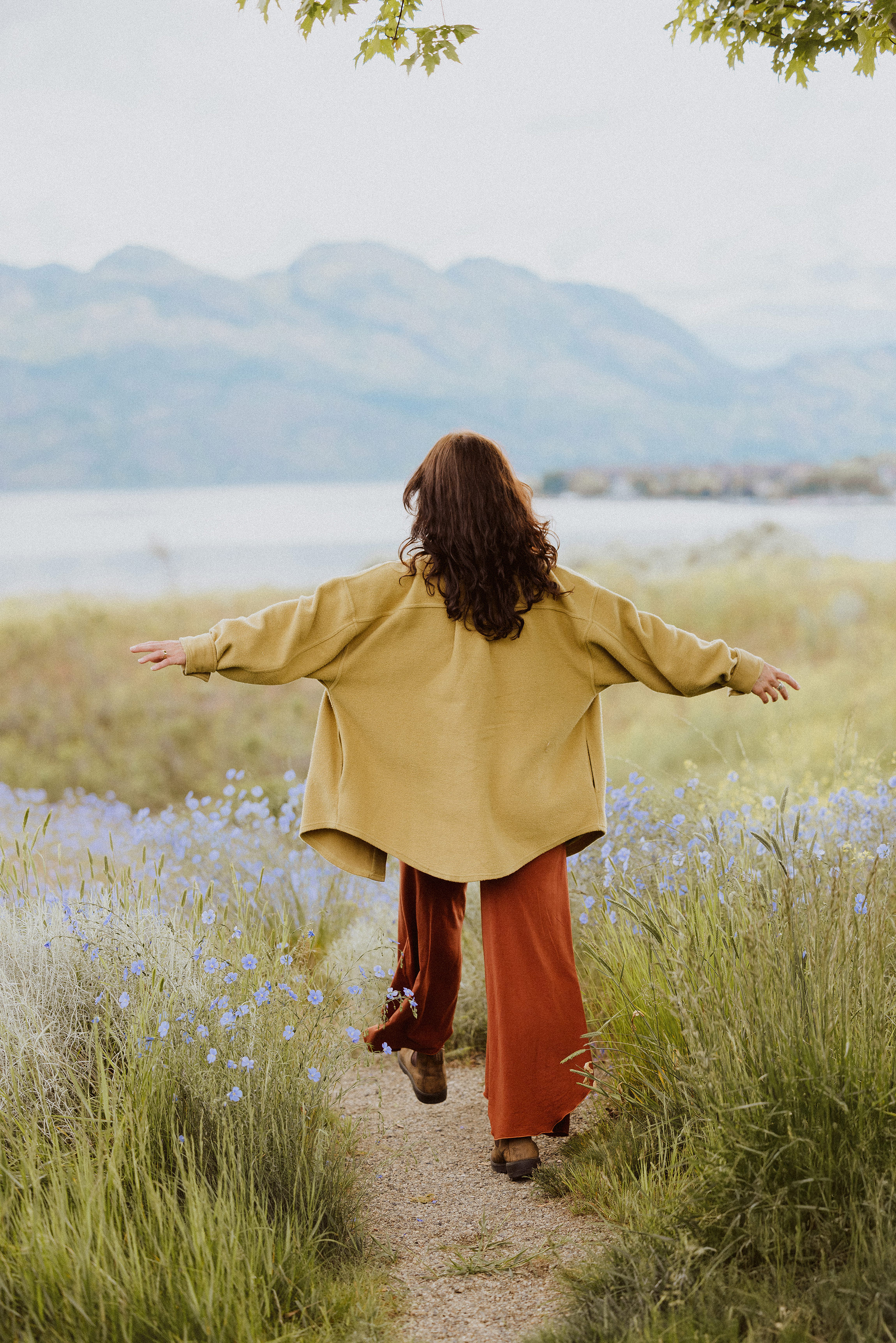 Woman walking along a wildflower path with arms outstretched toward a lake and mountains in the distance.