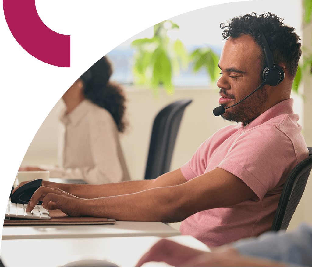 Customer support agent wearing a headset and pink shirt typing on a keyboard at a busy call center with a colleague behind.