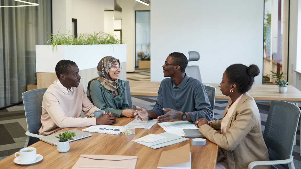 A team of four workers are sat at a boardroom table conversing.