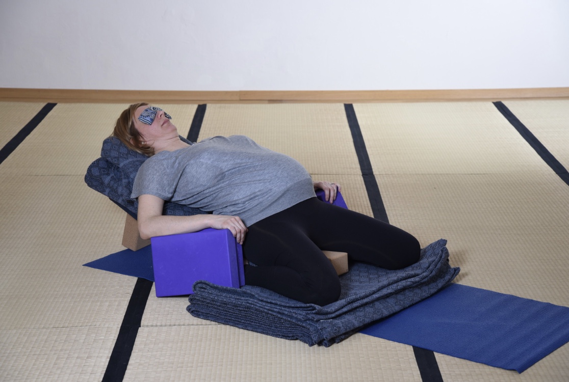 Femme enceinte en Supta Virasana, allongée sur un bolster, les bras posés sur des briques et un coussin sur les yeux