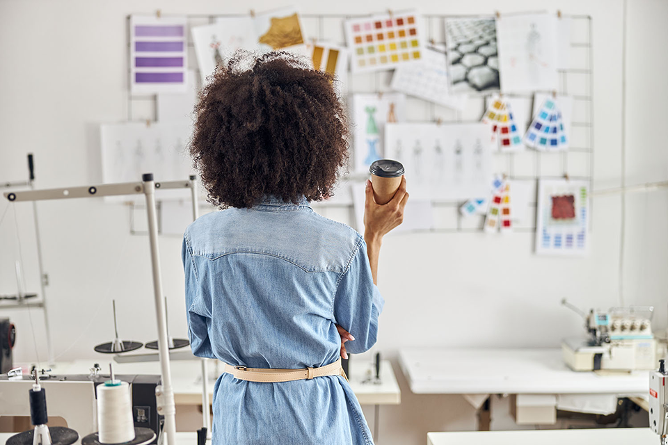 Back view of a fashion designer holding a coffee cup in a design studio, surrounded by sewing machines, pattern tools, sketches, and color swatches pinned to a wall.