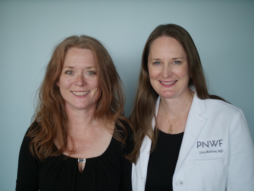 Two women smiling, both with long hair. The woman on the left is Stephanie and wearing a black shirt, the woman on the right is Lora and wearing her doctors coat.