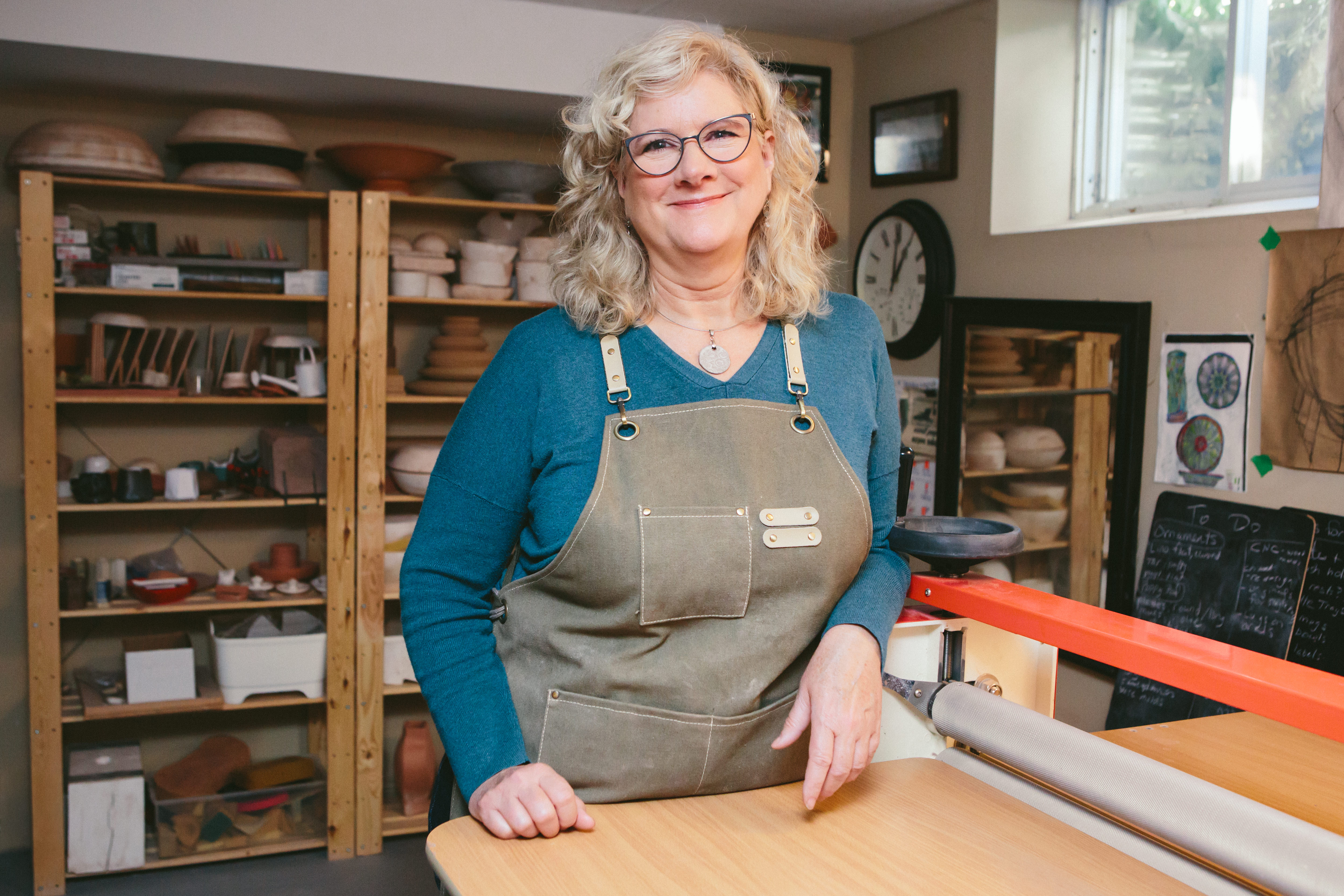 Image of Jacquie Blondin standing beside her slab roller with shelves behind her filled with examples and molds. 