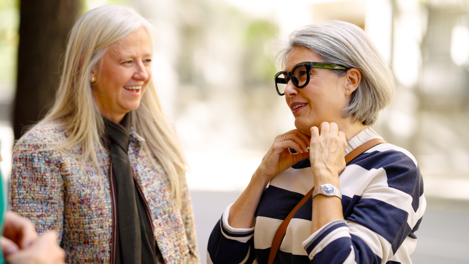Two older women standing outdoors in conversation, smiling and engaged with each other in a relaxed, friendly moment.