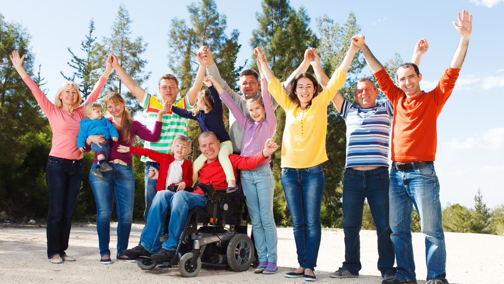 Diverse group of people standing together outdoors with arms raised, including a person in a wheelchair.